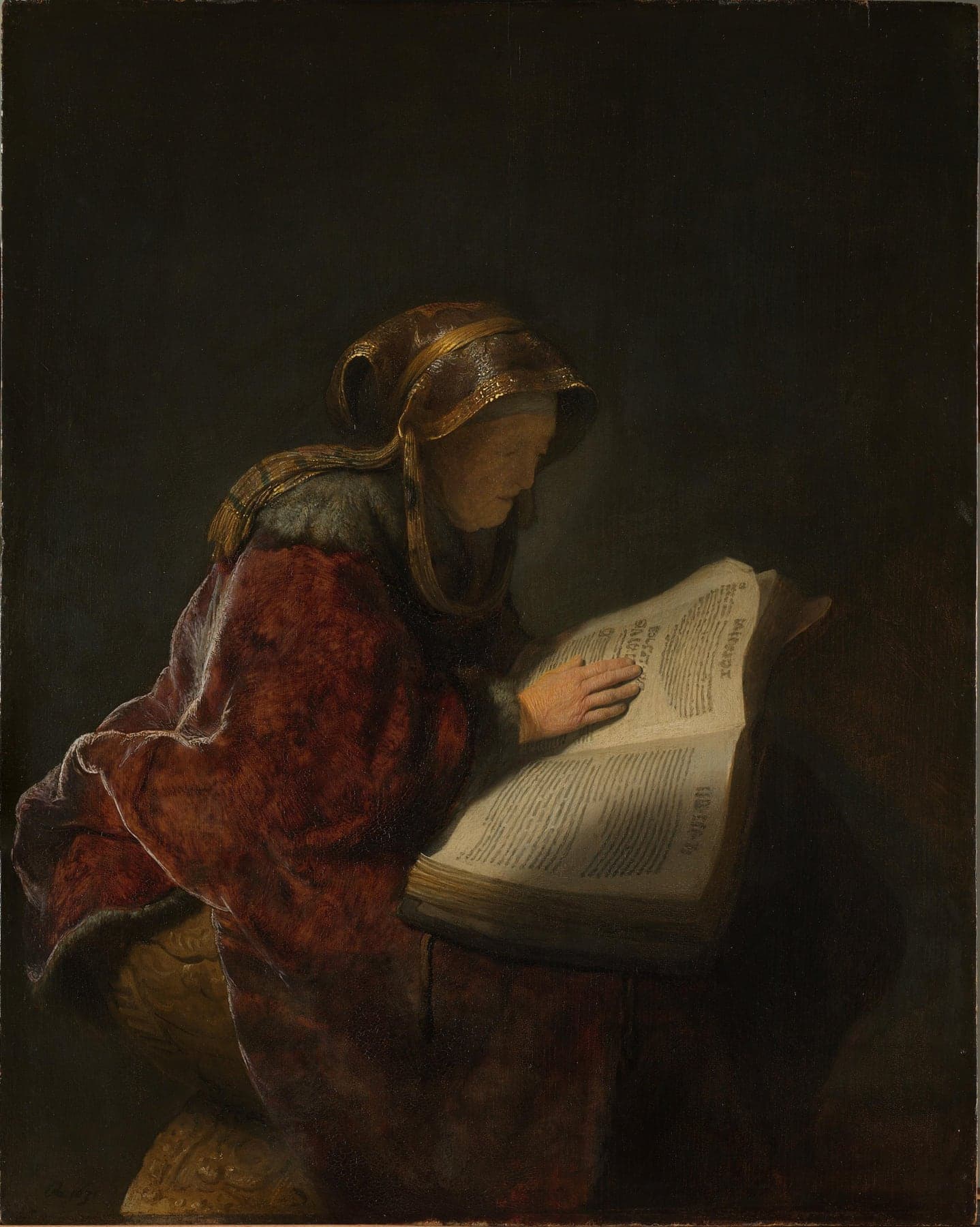 Rembrandt’s painting of an elderly woman in a red fur-trimmed robe and gold-embroidered headscarf, seated in profile in a dark room, one hand resting on the brightly lit pages of a large open book.
